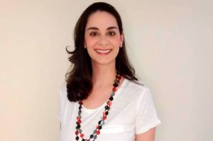 Mulher de cabelo escuro, camiseta branca e colar colorido sorri.