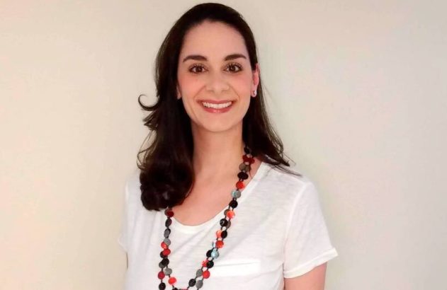 Mulher de cabelo escuro, camiseta branca e colar colorido sorri.