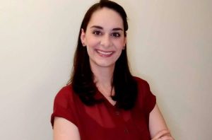 Retrato de mulher com cabelo escuro e camisa vinho, sorrindo para a câmera com os braços cruzados.
