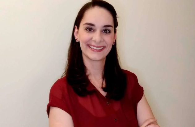Retrato de mulher com cabelo escuro e camisa vinho, sorrindo para a câmera com os braços cruzados.