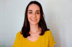 Retrato de mulher sorrindo, cabelo escuro, blusa amarela. Fundo de parede clara.