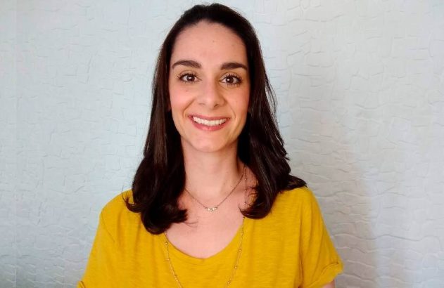 Retrato de mulher sorrindo, cabelo escuro, blusa amarela. Fundo de parede clara.