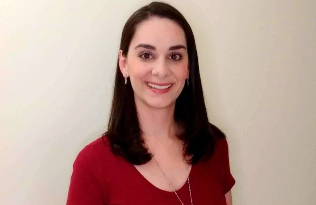 Mulher de cabelo escuro, camisa vermelha e colar, sorrindo para a câmera em fundo neutro.