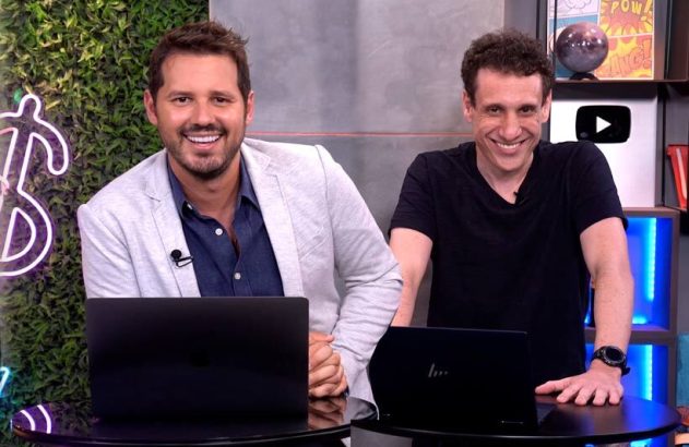 Dois apresentadores sorridentes sentados em um estúdio com laptops, um com parede verde e o outro com prateleiras.