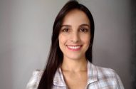 Retrato de mulher sorrindo, cabelo castanho escuro e camisa xadrez, em fundo neutro.