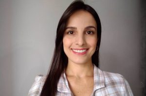 Retrato de mulher sorrindo, cabelo castanho escuro e camisa xadrez, em fundo neutro.