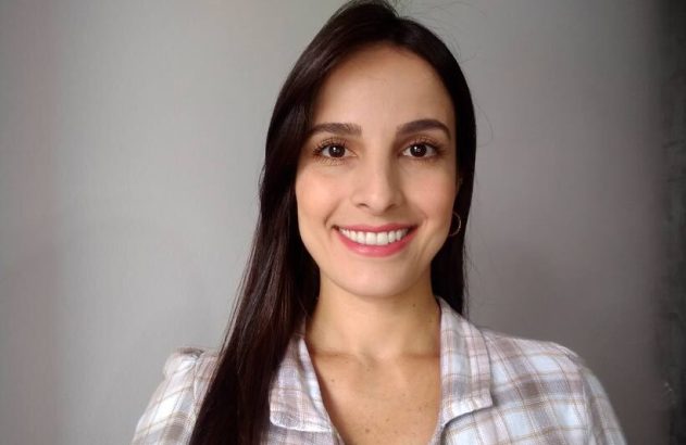 Retrato de mulher sorrindo, cabelo castanho escuro e camisa xadrez, em fundo neutro.