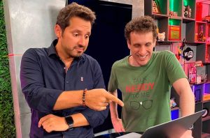 Dois homens em um estúdio: um aponta, surpreso, para um laptop, enquanto o outro ri usando camiseta "Talk Nerdy to Me".