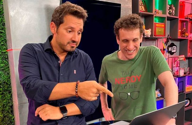 Dois homens em um estúdio: um aponta, surpreso, para um laptop, enquanto o outro ri usando camiseta "Talk Nerdy to Me".