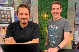 Dois homens sorridentes em um cenário de gravação. Um sentado com notebook, outro em pé. Ambos olham para a câmera.