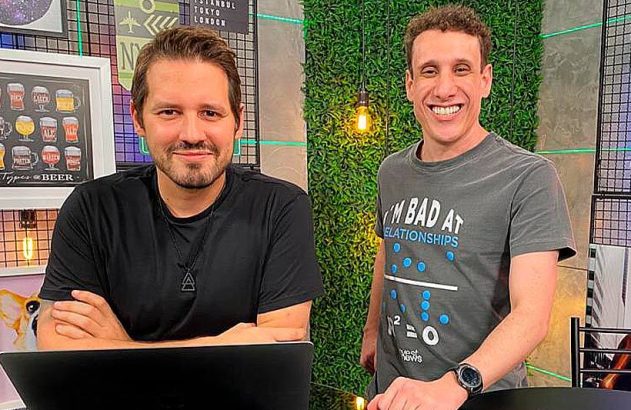 Dois homens sorridentes em um cenário de gravação. Um sentado com notebook, outro em pé. Ambos olham para a câmera.