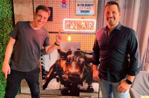 Dois homens sorrindo posam ao lado de uma estátua de minotauro iluminada. O da esquerda aponta para a figura.