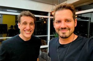 Dois homens sorrindo em um ambiente interno com grandes janelas e vista noturna de uma cidade iluminada.