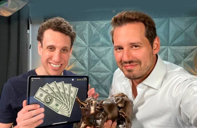 Dois homens sorrindo. Um exibe um tablet com dinheiro, o outro segura um troféu.