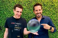 Dois homens sorrindo, um segura uma esfera de cristal e o outro aponta para ela. Fundo verde.