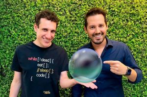 Dois homens sorrindo, um segura uma esfera de cristal e o outro aponta para ela. Fundo verde.