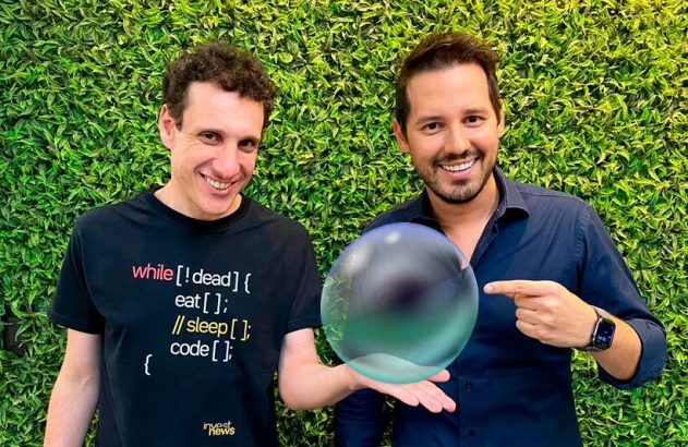 Dois homens sorrindo, um segura uma esfera de cristal e o outro aponta para ela. Fundo verde.