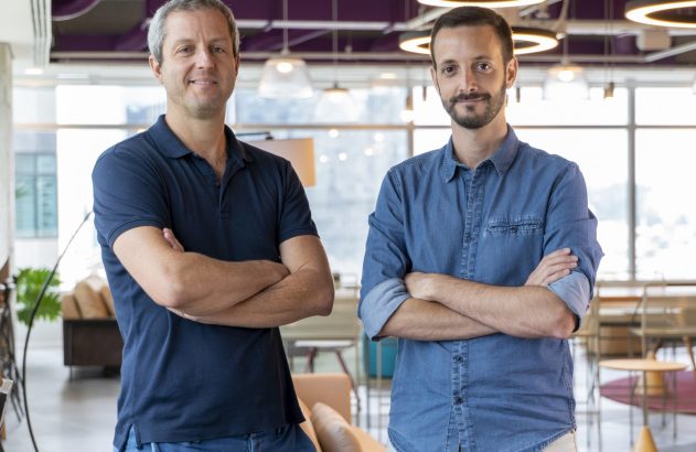 Dois homens de braços cruzados olhando para a câmera em um escritório moderno.