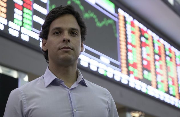 Homem de camisa clara com painel de cotações de bolsa ao fundo.