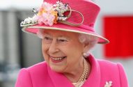 Rainha Elizabeth II sorrindo, com chapéu pink floral, casaco combinando e colar de pérolas.