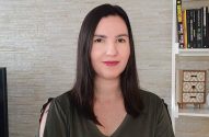 Mulher de cabelo escuro e blusa verde sorrindo para a câmera. Fundo de parede clara e estante de livros.