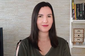 Mulher de cabelo escuro e blusa verde sorrindo para a câmera. Fundo de parede clara e estante de livros.