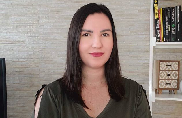 Mulher de cabelo escuro e blusa verde sorrindo para a câmera. Fundo de parede clara e estante de livros.