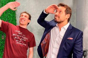 Dois homens sorrindo, um de camiseta e um de terno, olham à distância protegendo os olhos com as mãos.