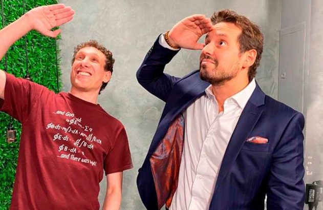 Dois homens sorrindo, um de camiseta e um de terno, olham à distância protegendo os olhos com as mãos.