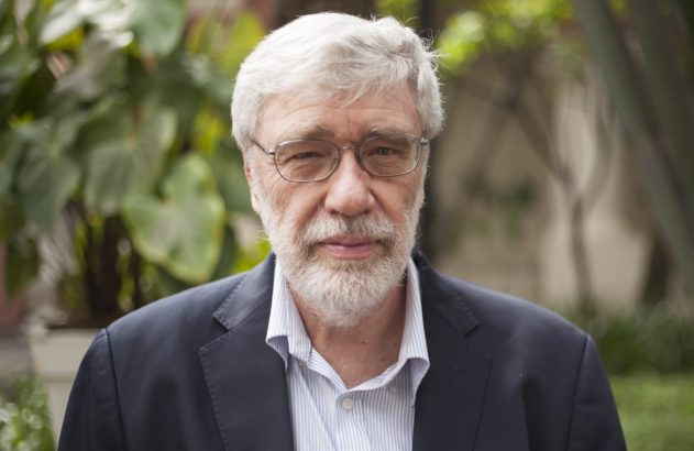 Retrato de um homem com cabelo e barba grisalhos e óculos, vestindo um casaco escuro, olhando para frente.