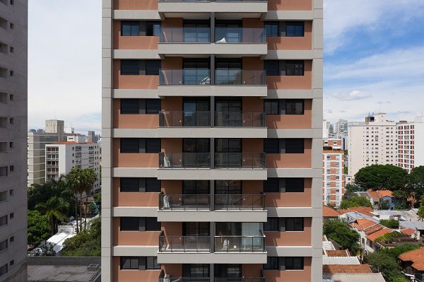 Prédio residencial de andares altos, fachada salmão e cinza, varandas com vidro, em área urbana com casas e edifícios.