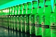 Fileira de garrafas Heineken verdes, com rótulos visíveis, sob luz esverdeada e reflexos.