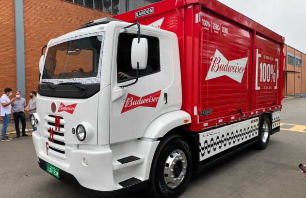 Caminhão elétrico e solar Budweiser branco/vermelho em apresentação, com pessoas.