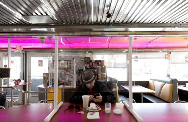 Homem de chapéu usa celular em mesa de lanchonete com barreira plástica e iluminação neon rosa.