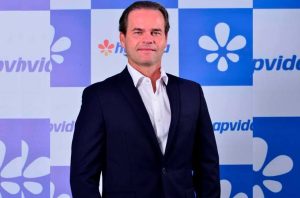 Homem de terno e camisa branca, em pé, em frente a um fundo azul e branco com o logo da Hapvida.