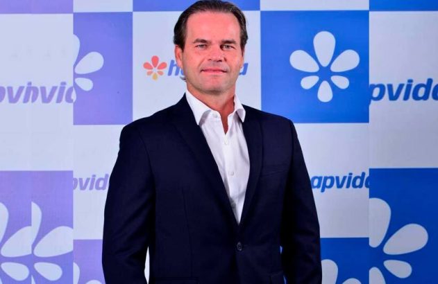 Homem de terno e camisa branca, em pé, em frente a um fundo azul e branco com o logo da Hapvida.