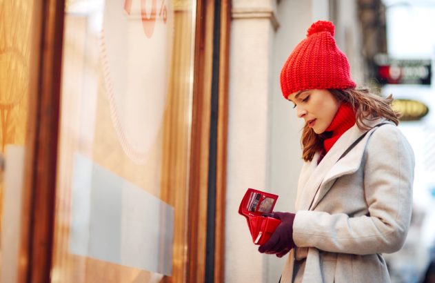 Mulher de gorro e cachecol vermelho mexe em sua carteira em frente a uma vitrine de loja.