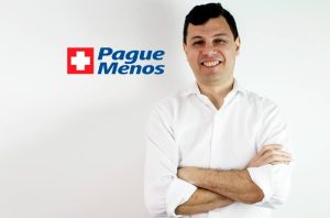 Homem sorridente de camisa branca, braços cruzados, com logo Pague Menos e cruz vermelha.