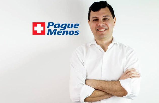Homem sorridente de camisa branca, braços cruzados, com logo Pague Menos e cruz vermelha.