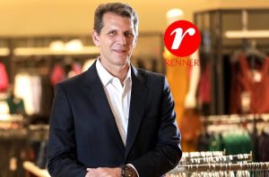Homem de terno sorrindo em loja de roupas. Logotipo Renner no canto superior direito.