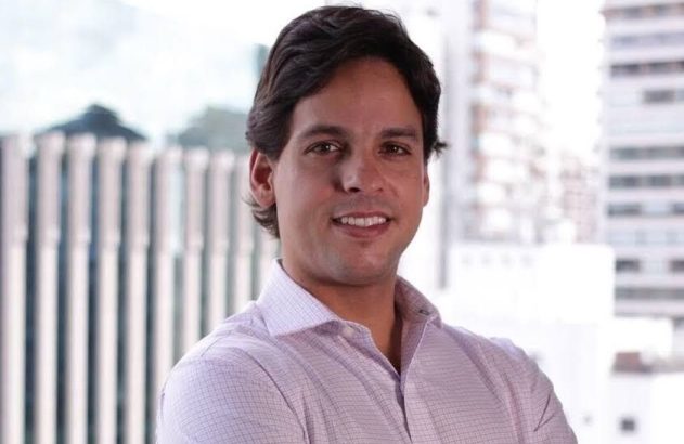 Retrato de homem sorrindo, camisa social e braços cruzados, com fundo de prédios da cidade.