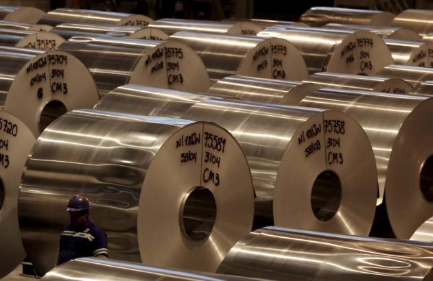 Fileiras de grandes bobinas de metal brilhante em armazém industrial, com operário de capacete azul.
