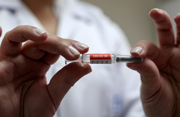 Mãos segurando uma ampola de vidro com o rótulo vermelho "COV-02-IB". Produto de pesquisa médica.