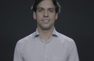 Homem sorrindo, de camisa, em fundo escuro.