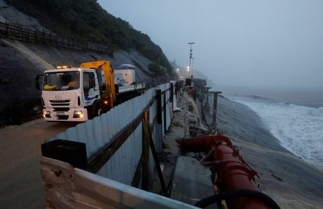 Caminhão da CEDAE em estrada costeira molhada. Ondas na margem com tubulação e cercas de construção.