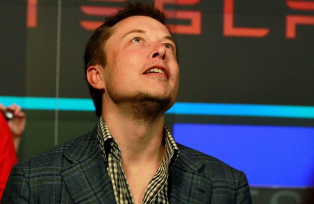 Elon Musk está com um blazer preto olhando para cima em frente ao logo da Tesla