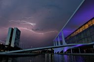 Raios no céu noturno sobre o Congresso Nacional e Palácio Itamaraty roxo, com reflexos na água.