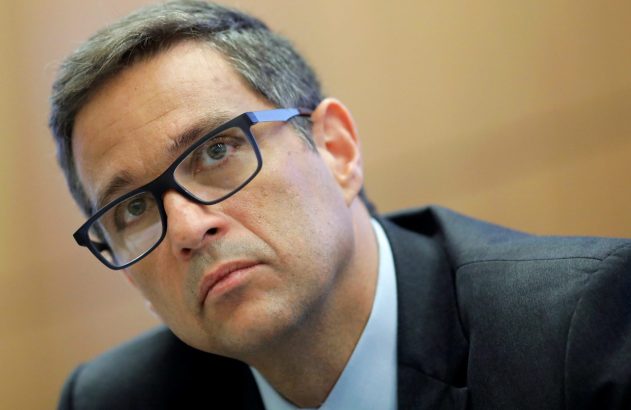 Homem de terno escuro e óculos, com cabelo grisalho, olhando fixamente para cima com expressão séria.