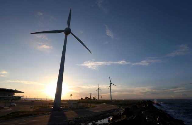Turbinas eólicas em silhueta sob céu crepuscular ao lado de rochas e mar agitado.