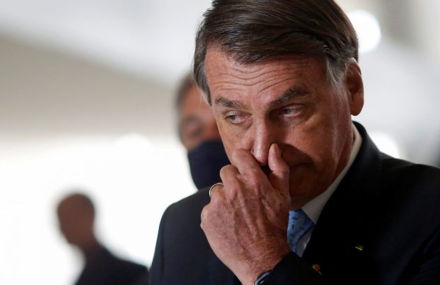 Jair Bolsonaro, de terno e gravata, olha para baixo com a mão no nariz, expressão pensativa.